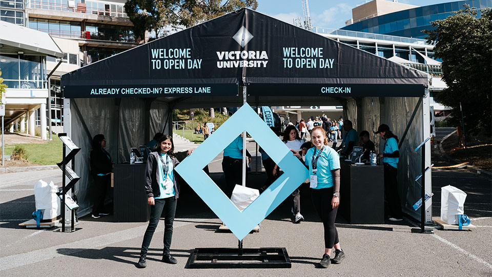 Two students stand either side of a replica of the VU blue branded diamond at Open Day, 2023, Footscray Park.