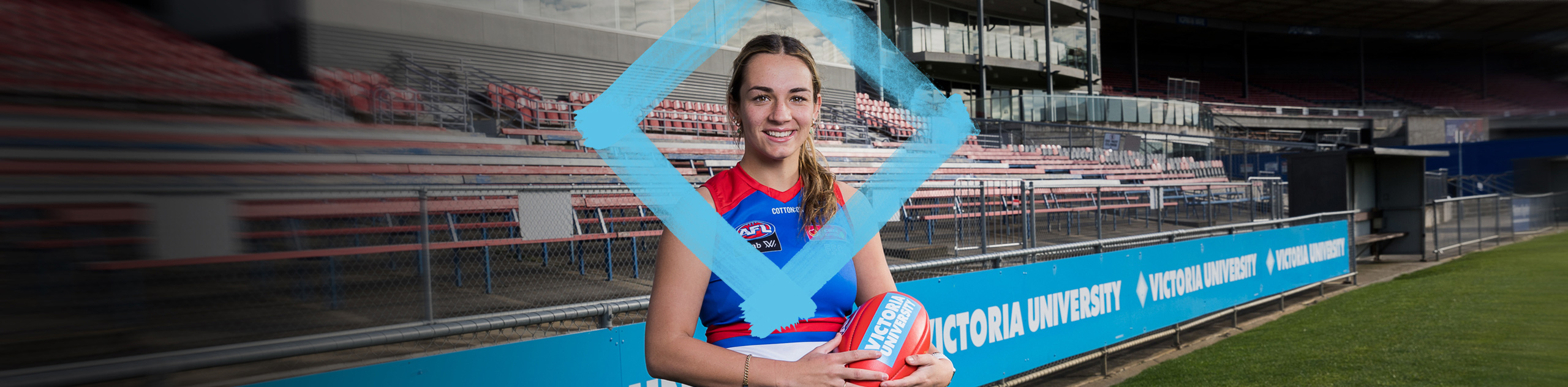 A young woman in a football uniform holds an oval ball that reads 'Victoria University, on a sport field with Victoria University banners