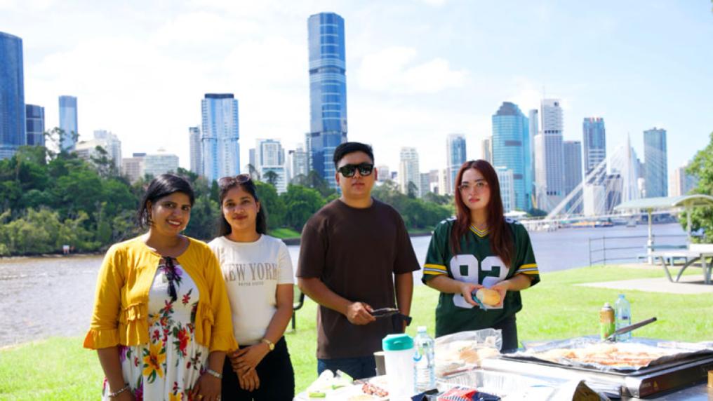Four students standing outside in front of a barbeque. Behind them is the Brisbane river and city buildings.