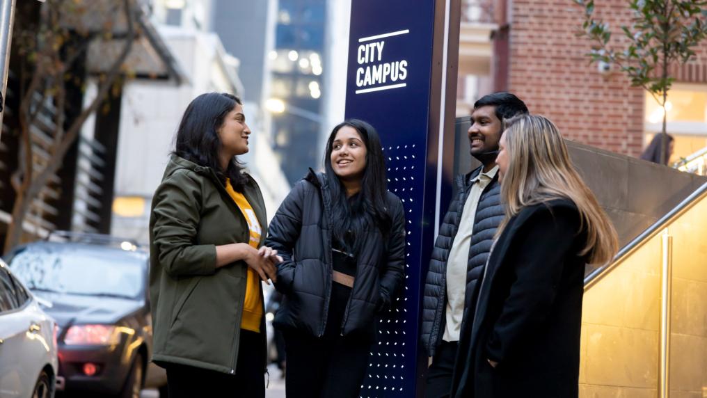 4 students standing outside VU's City Campus and smiling. The city landscape is behind them, with cars parked on the street.