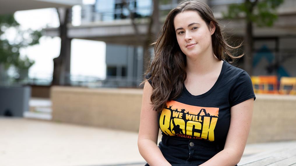 A young student with long hair and a black 'rock' t-shirt sits in a campus courtyard